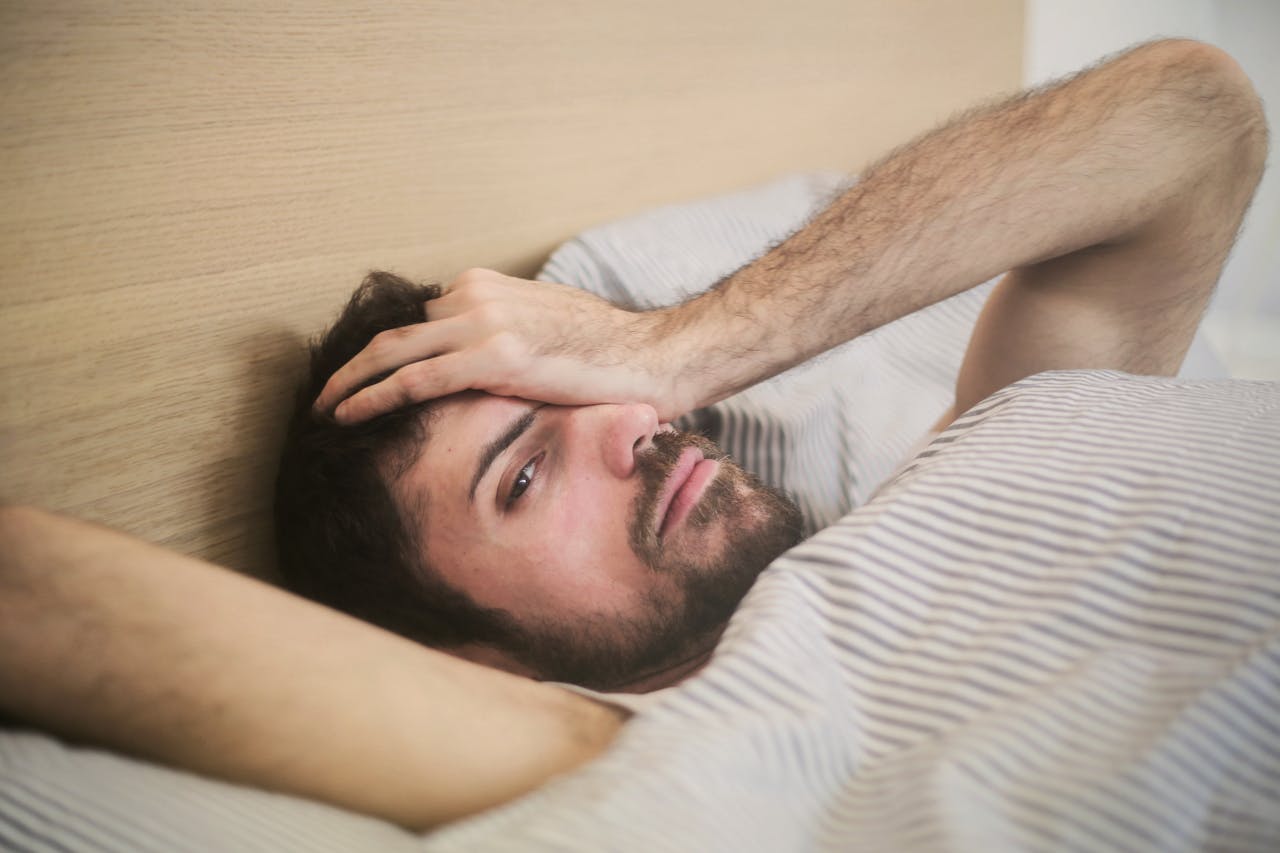 Man lying in bed looking fatigued and holding his head, experiencing symptoms of tiredness or low energy.