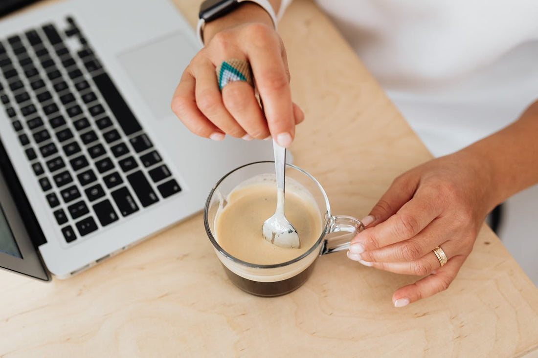 Woman stirring coffee at a desk beside a laptop.