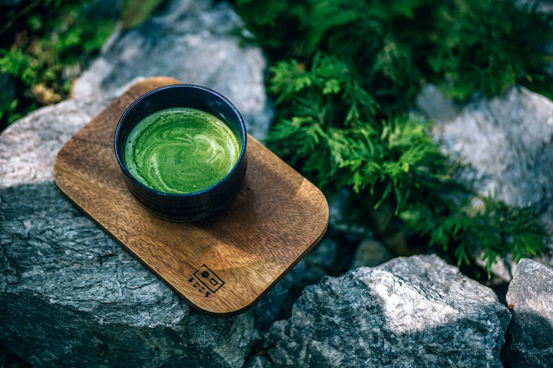 A bowl of green tea sits on a wooden tray placed on rocky terrain.
