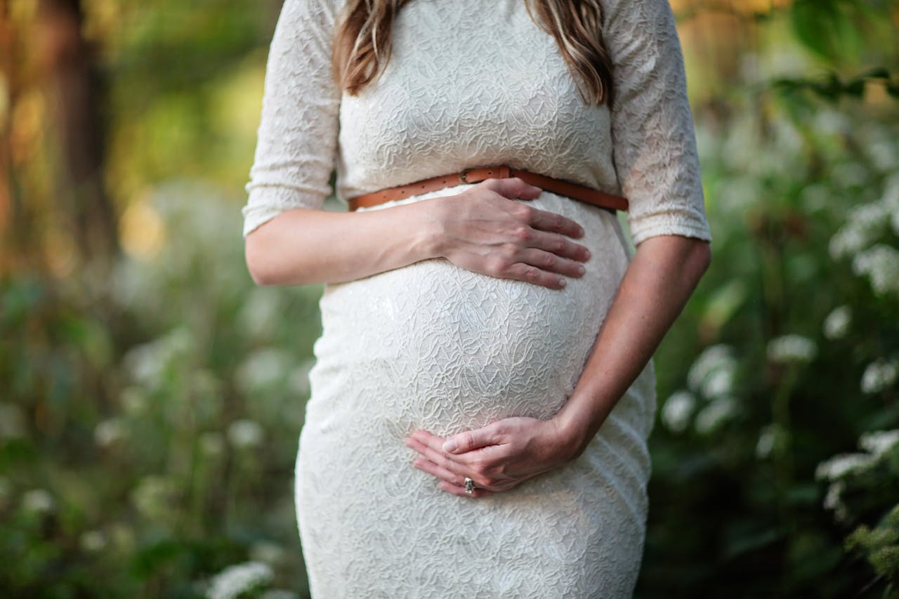 A pregnant woman in a white lace dress holding her belly with both hands.
