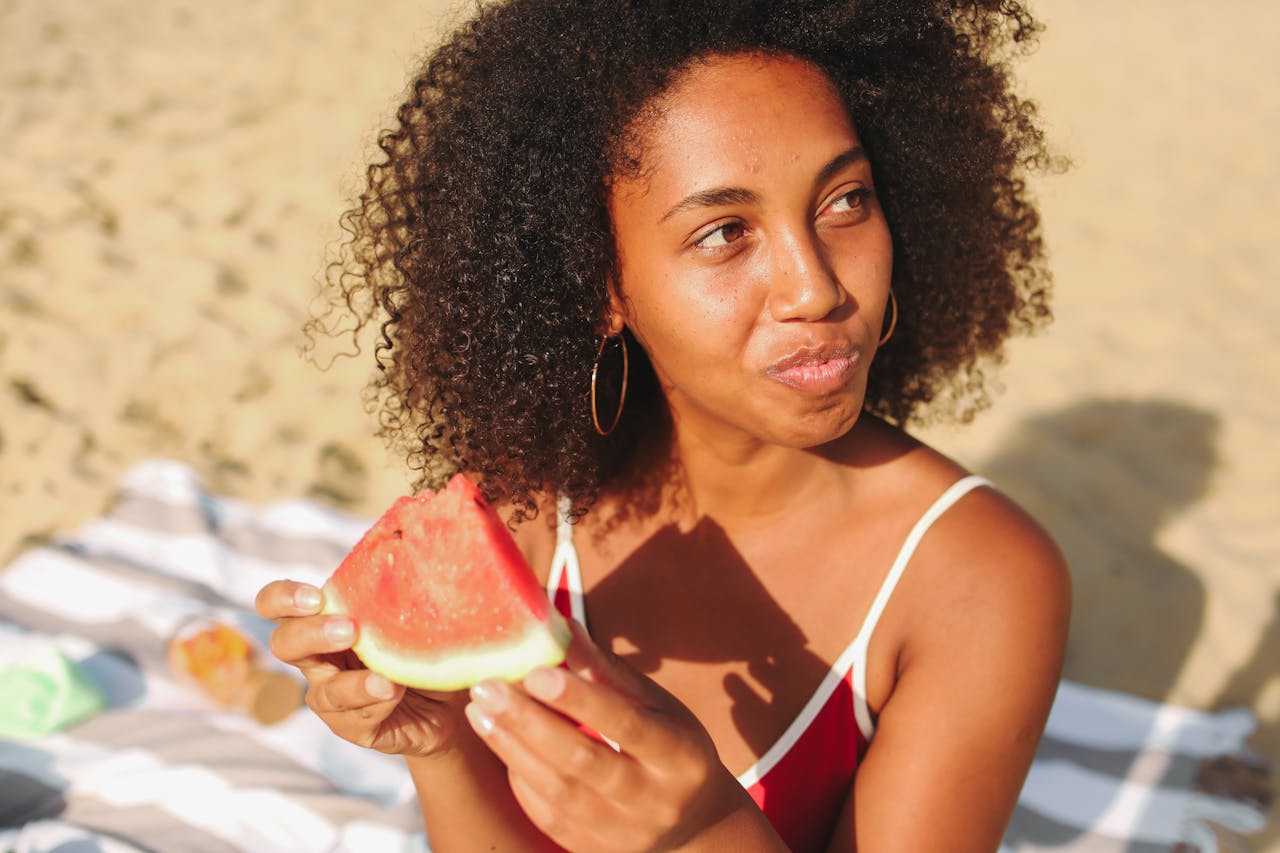 A woman with curly hair enjoying a slice of watermelon while at the beach.