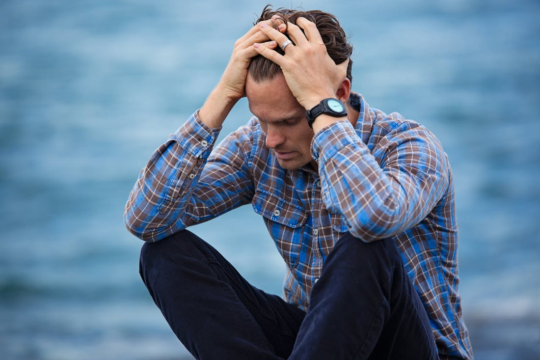 Man sitting outdoors near the water, holding his head in frustration, showing signs of emotional distress.
