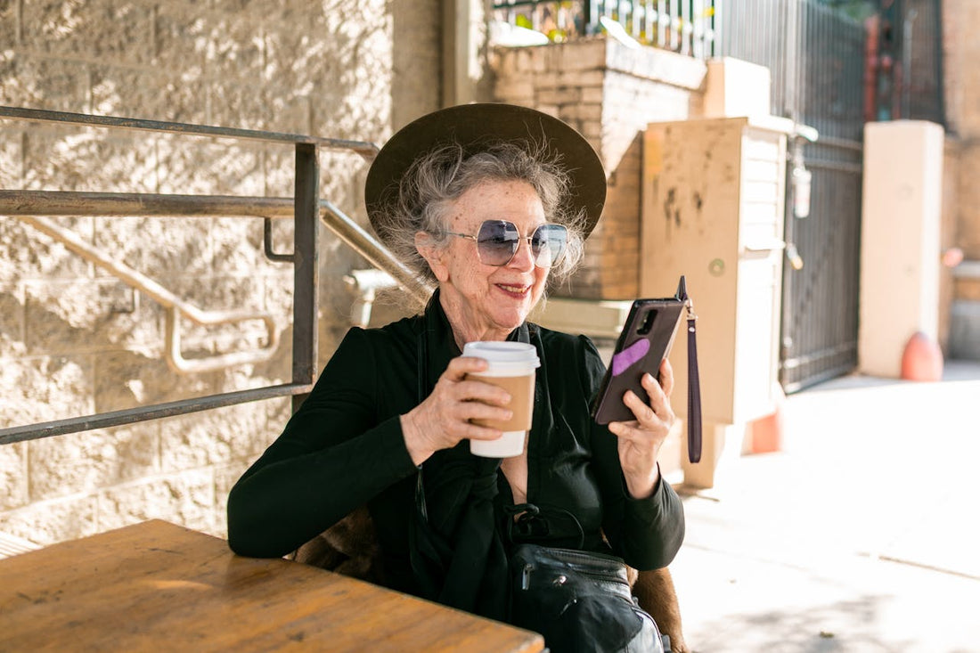 Older woman sitting at outdoor café, smiling as she looks at a smartphone while holding a cup of coffee